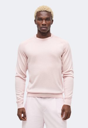 Schwarzer Mann mit kurz gebleichtem Haar, der einen blassrosa Langarmpullover und passende hellrosa Hose trägt, steht vor einem weißen Hintergrund.