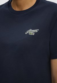 Μπλε ναυτικό t-shirt με στρογγυλή λαιμόκοψη με το λογότυπο Lacoste που περιλαμβάνει πράσινο κροκόδειλο και κείμενο με καλλιγραφία στο αριστερό στήθος, φορεμένο από ένα άτομο.