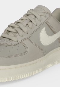Ανοιχτόγρινο παπούτσι Nike με λευκό λογότυπο swoosh, μπεζ κορδόνια και υφασμένο λευκό πέλμα, εμφανίζεται σε κοντινή λήψη σε μονόχρωμο φόντο.