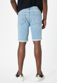 Lysblå denimshorts med opkrøllede manchetter, med baglommer og et lædermærke ved taljen. Model iført hvide og mørke sneakers.