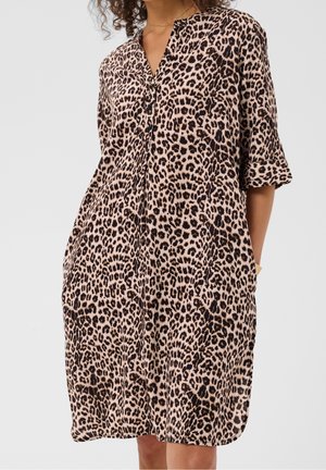 Kvinde iført en løs leopardmønstret kjole med ærmer til midt på armen og knaplukket V-hals, stående med hænderne i lommerne.