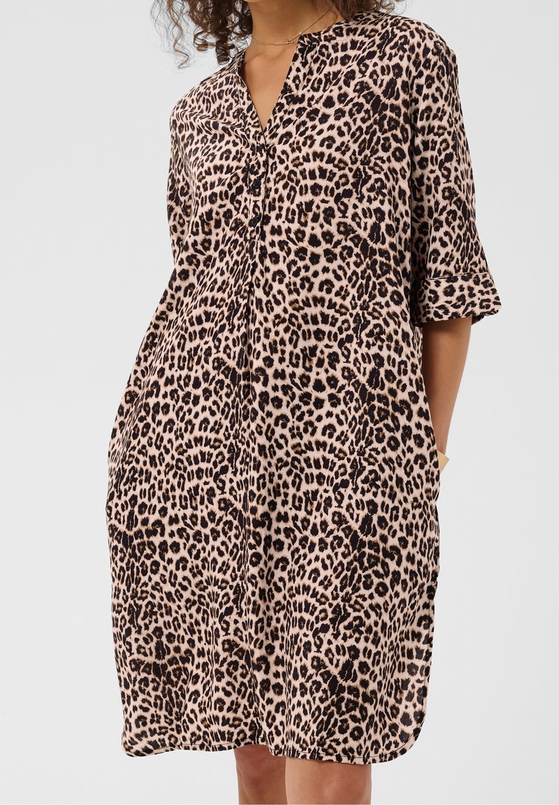 Femme portant une robe ample à imprimé léopard avec des manches mi-longues et un col en V boutonné, debout les mains dans les poches.