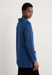 Kobieta z krótkimi włosami, ubrana w niebieską, w paski, długą koszulę z długim rękawem, patrzy w dół, zwrócona bokiem, z lewą ręką swobodnie opartą na boku.