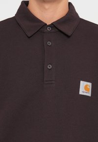 Dark brown polo shirt made of textured cotton, featuring a buttoned collar and a rectangular Carhartt logo patch with orange accents.