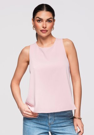 Donna con capelli scuri raccolti in una coda di cavallo che indossa orecchini a cerchio dorati, un top rosa pallido senza maniche e jeans blu su uno sfondo chiaro uniforme.