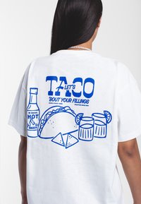 Bílý tričko s modrým grafickým designem zobrazujícím taco, chipsy, dva nápoje a text "TACO LET'S 'BOUT YOUR FILLINGS". Pohodlný střih.