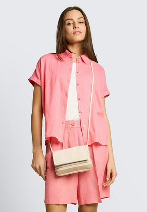 Jonge vrouw die een loszittend roze shirt met korte mouwen draagt, bijpassende korte broek, een wit topje en een beige schoudertas tegen een effen achtergrond.