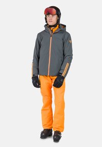Grau wasserdichte Jacke mit orangen Akzenten, orange wasserdichte Hose, schwarze Handschuhe und Skistiefel. Modell steht vor einem weißen Hintergrund.