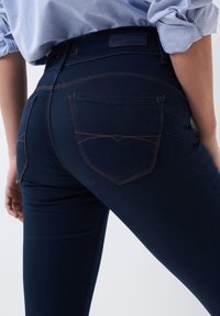 Ciemnoniebieskie dżinsy o dopasowanym kroju, z pomarańczowymi szwami i dwiema tylnymi kieszeniami. W pasie znajduje się metka z logo marki.