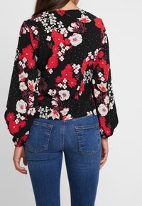 Blusa floreale con sfondo nero, decorata con grandi fiori rossi e bianchi; maniche lunghe e vestibilità morbida. Indossata con pantaloni in denim blu.