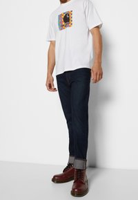 Hombre vestido con una camiseta gráfica blanca, jeans oscuros arremangados y botas de cuero rojo con cordones, de pie contra un fondo blanco.