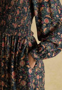 Kleid mit floralem Muster, mit gerafftem Mieder, langen Ärmeln und Taschen. Dunkel marineblauer Hintergrund mit bunten Blumen- und Paisleymustern.