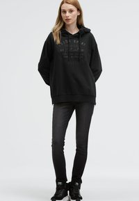 Black hooded sweatshirt with ribbed hem and cuffs features a printed design in front. Paired with dark skinny jeans and black sneakers.