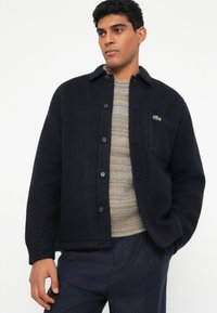 Marineblauwe fleece overhemdjas met knoopsluiting, borstzak en geborduurd logo. Draagt over een meerkleurige trui en gecombineerd met een donkere broek.