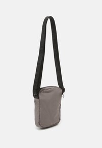 Gray fabric crossbody bag with a zip closure, adjustable black strap featuring logo, and soft, textured surface. Compact rectangular shape.