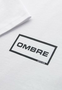 Hvidt stof med et sort rektangulært logo, der indeholder ordet "OMBRE" med fede store bogstaver og mindre "DESIGN" tekst nederst til højre.