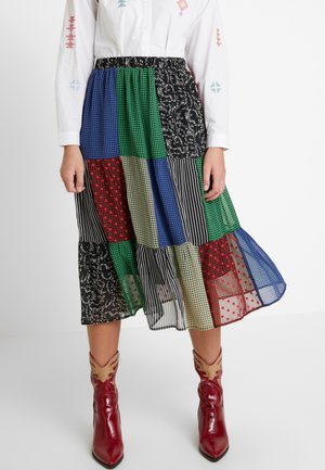 Mujer con falda midi patchwork con estampados y colores mezclados, camisa blanca de manga larga y botas de vaquero rojas puntiagudas.