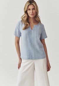 Blusa di lino azzurro chiaro con maniche corte, scollo a V e pieghe verticali sulla parte frontale. Abbinata a pantaloni bianchi larghi. Texture liscia.