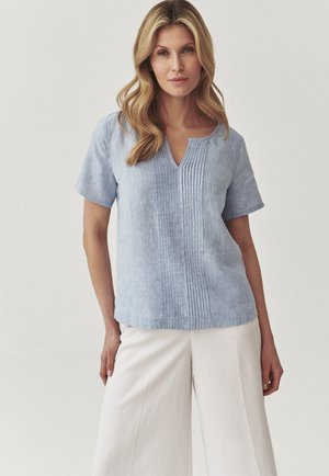 Blusa di lino azzurro chiaro con maniche corte, scollo a V e pieghe verticali sulla parte frontale. Abbinata a pantaloni bianchi larghi. Texture liscia.