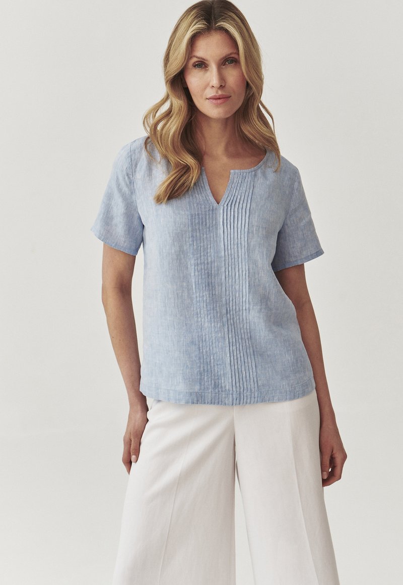 Blusa di lino azzurro chiaro con maniche corte, scollo a V e pieghe verticali sulla parte frontale. Abbinata a pantaloni bianchi larghi. Texture liscia.