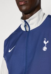 Giacca Nike navy e bianca con colletto alto, chiusura a zip e logo degli Spurs sul lato sinistro del petto. Presenta una trama di tessuto liscia.
