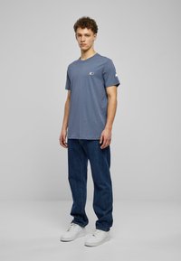 Gray cotton T-shirt with short sleeves, featuring a small logo on the left chest. Paired with dark blue jeans and white sneakers.