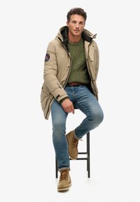 Béžová péřová bunda s černým podšívkou, zelený strukturovaný svetr, modré džíny a hnědé boty. Model sedící na černém stoličce.