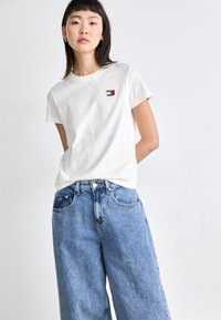 Witte katoenen T-shirt met korte mouwen, voorzien van een klein rood en blauw logo-opdruk op de borst. Gepaarde met losse, lichtblauwe denim jeans.