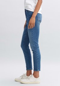 Žena v modrých džínech s mírným pasem a štíhlým střihem, obutá v bílých teniskách, stojící na jednobarevném světle šedém pozadí.