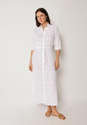 Frau mit dunklen Haaren trägt ein weißes besticktes langes Hemdblusenkleid mit 3/4-Ärmeln, gebundener Taille und beigen flachen Sandalen, steht an einer schlichten Wand.