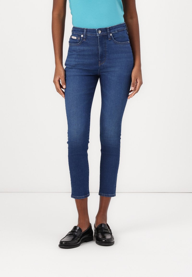 Jeans de tiro alto azules con un ajuste slim, de longitud recortada, cinco bolsillos y detalles de costura sutiles. Combinados con mocasines negros.