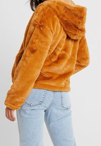 Hnědo-červený umělý kožešinový hoodie se strukturovaným povrchem, velkou kapucí a volným střihem, kombinovaný se světle modrými džíny.