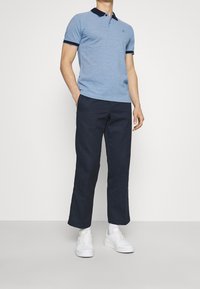 Polo bleu clair avec col et poignets bleu marine, associé à un pantalon bleu marine foncé. Des baskets blanches complètent la tenue. Design simple et épuré.