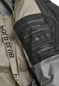 Beige ytterjacka med en svart lapp med vit text, en strukturerad mönster och ett märkesband. Detaljer inkluderar ikoner för vattentålighet.