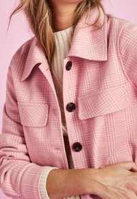 Roze geruite jas met een kraag, twee zakken aan de voorkant en donkere knopen. Zachte textuur, gedragen over een lichtgekleurde trui.