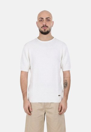 Uomo con barba e tatuaggio che indossa un maglione bianco a maniche corte con texture e pantaloncini beige, in piedi davanti a uno sfondo chiaro e uniforme.