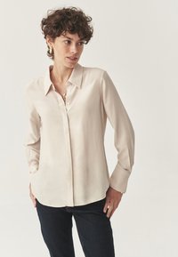 Camicia button-up beige chiaro con colletto, maniche lunghe e una texture morbida e setosa. Abbinata a jeans scuri con tasche laterali.