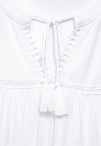 Vit blus med rund halsringning, prydd med virkade spetsdetaljer och ett knytband. Två vita fransar hänger från halsringningen. Lättviktsmaterial.