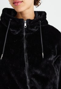 Zwarte nepbont hoodie met een rits aan de voorkant, voorzien van een hoge kraag en een trekkoord. De capuchon heeft een zachte textuur en een subtiele glans.