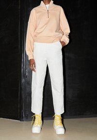 Vaalean persikanvärinen vetoketjullinen huppari, jossa on pallopaisutetut hihat, yhdistetty valkoisiin lyhyisiin housuihin ja tukevat valkoiset tennarit, joissa on keltaiset vaijerit.