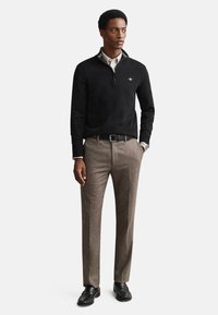 Crni džemper s dugim rukavima i patentnim zatvaračem na ovratniku, uparen s bež teksturiranim hlačama i crnim cipelama. Na džemperu je vidljiv diskretni logo.