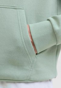 Lichtgroene sweatshirt met een zachte textuur, voorzien van een kangoeroezak en geribde manchetten. Gedetailleerde stiksels zichtbaar langs de naden.