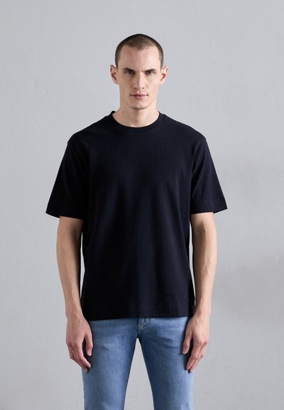 T-shirt noir à texture, à manches courtes et col rond, associé à un jean bleu clair, porté par un mannequin sur un fond neutre.