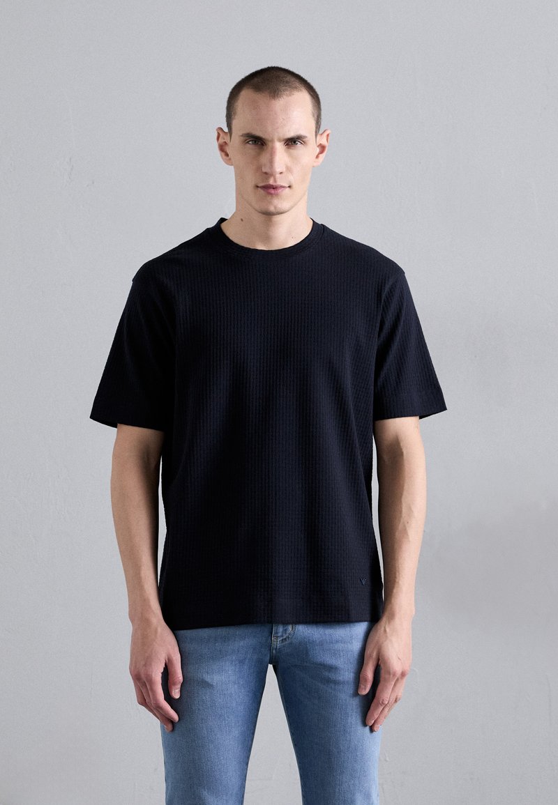 Camiseta preta texturizada de manga curta com decote redondo, combinada com jeans azul claro, usada por um modelo contra um fundo neutro.