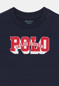 Marinblå bomullströja med en djärv röd och vit grafik som säger "POLO RALPH LAUREN." Den har en rundad nacke och tryckknappar.