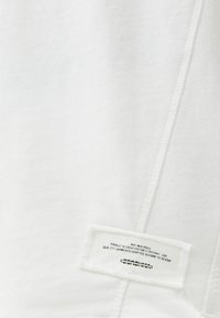 Bílá mikina s texturovanou tkaninou, která obsahuje prošitou ševovou detal. Na boku je umístěná malá péče štítek s pokyny pro údržbu.