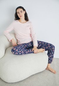 Ljusrosa långärmad topp med en ficka, matchad med marinblå byxor med blommönster, sittande på en texturerad krämfärgad soffa.