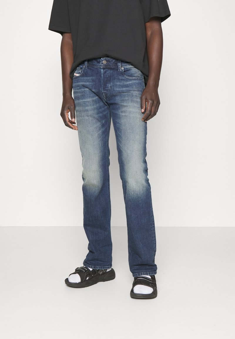 Personne portant un jean bleu délavé, des sandales noires avec des chaussettes blanches et un t-shirt noir ample sur un fond clair uni.
