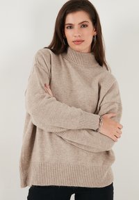 Beige kudumütsiga kampsun, millel on kõrge soonikuga krae, lõdva istuvuse ja soonikuga alläär. Pehme tekstuur, kukkunud õlad ja pikad varrukad.