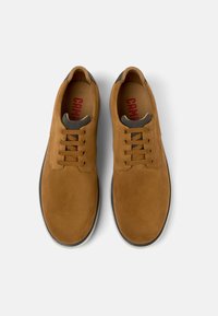 Pantofi din suede maro, cu o textură netedă, având șase gauri pentru șireturi, șireturi maro și un căptușel gri în jurul gleznei. Inscripție roșie a brandului în interior.